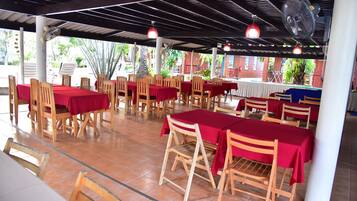 An outdoor restaurant with wooden tables and chairs, covered by a roof with fans and hanging lights.