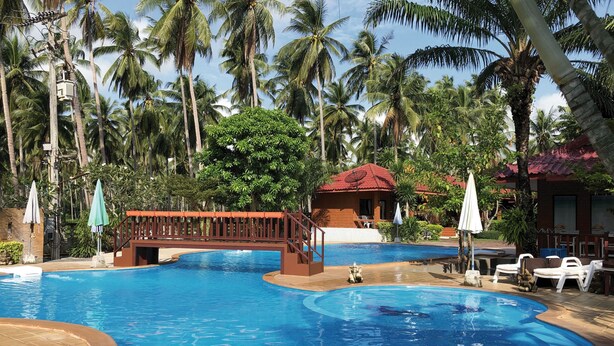 A swimming pool with a bridge, surrounded by palm trees and a red-roofed building. - Bankrut Resort (Bang Saphan)