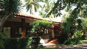 A building with a red-tiled roof, a stone facade, and a garden with various plants and a large flowering bush. - Bankrut Resort (Bang Saphan)