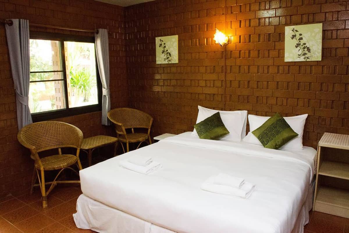 A hotel room with a brick wall, a bed with white linens, two green pillows, a nightstand, a wicker chair, and a window with curtains.