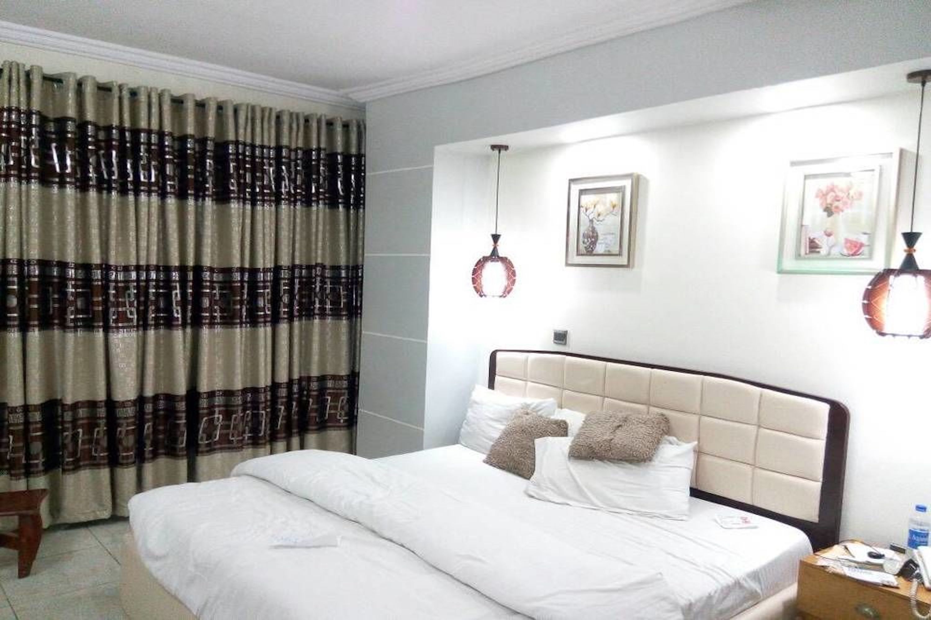 A neatly made bed with white linens and two decorative pillows, a bedside table with a phone and a bottle, and a wall-mounted light fixture.