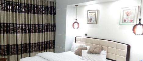 A neatly made bed with white linens and two decorative pillows, a bedside table with a phone and a bottle, and a wall-mounted light fixture.