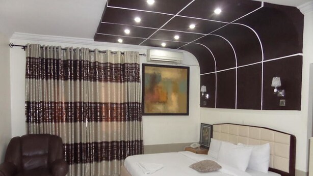 A hotel room with a bed, a chair, a painting, and a ceiling with recessed lighting. - Prumssy Hotel (Lekki)