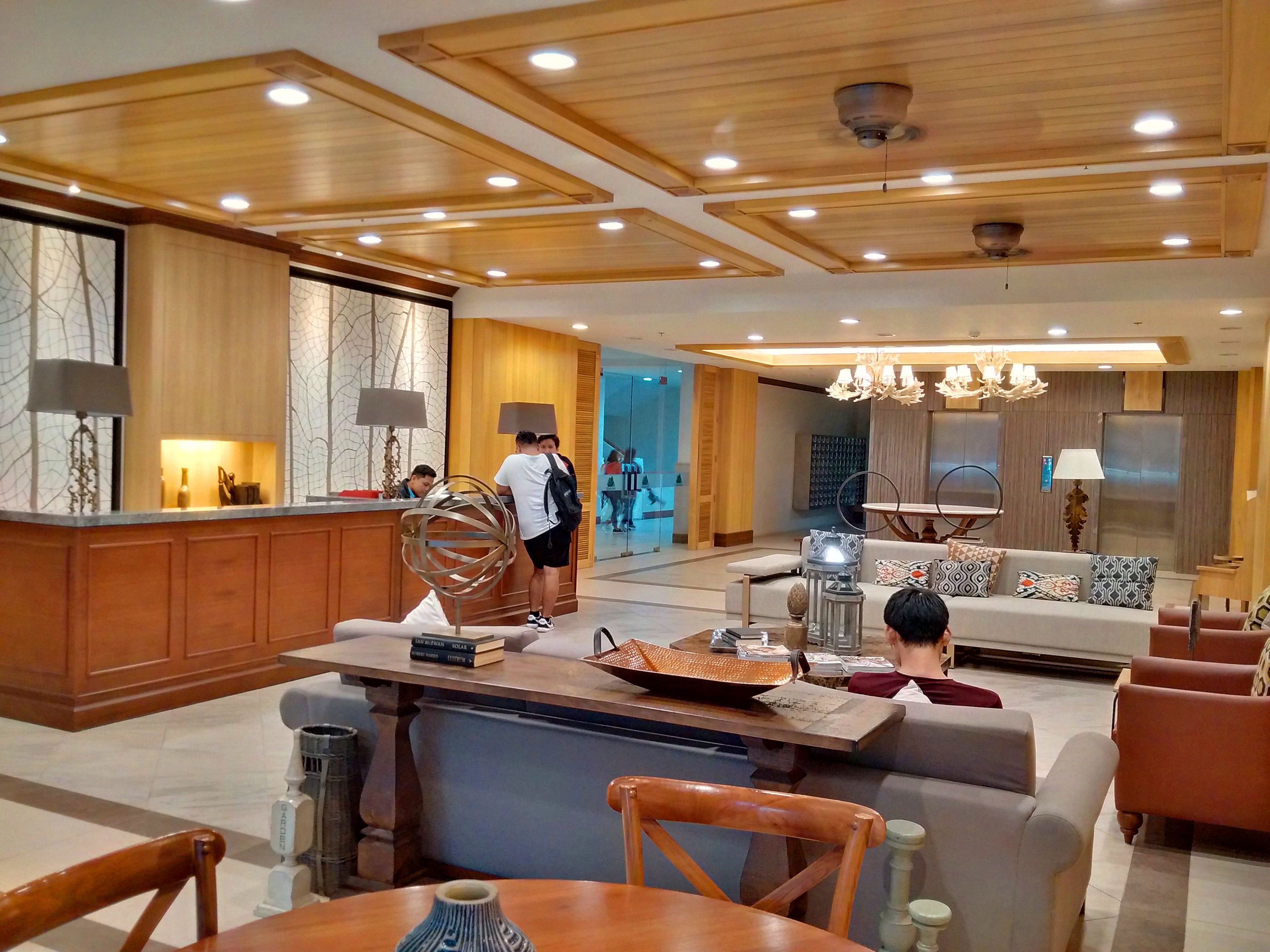 Een moderne hotellobby met houten balken in het plafond, een receptiebalie, een zithoek met banken en een eettafel, en een kroonluchter.