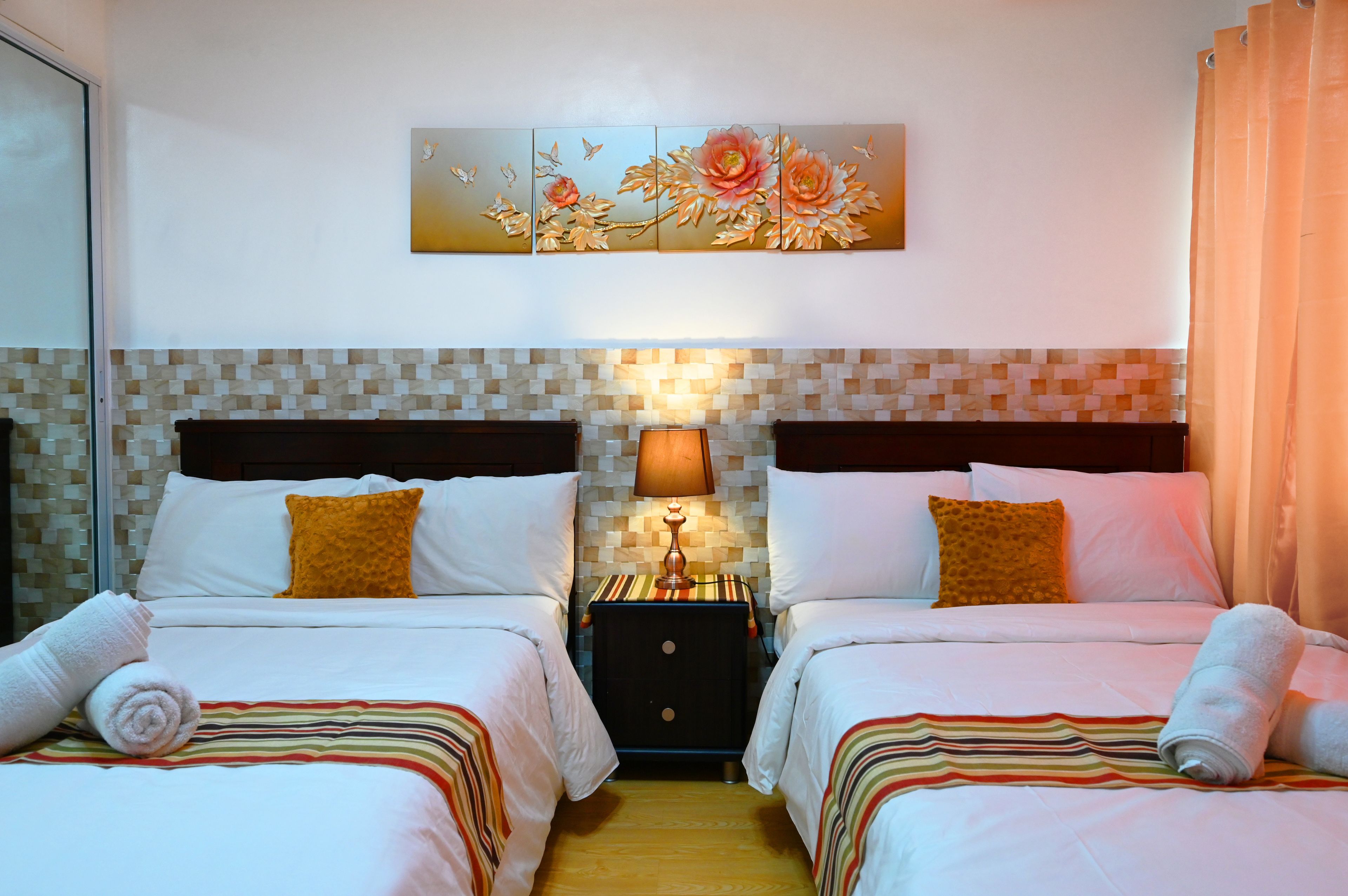 A hotel room with two beds, a nightstand with a lamp, a decorative wall piece, and a tiled backsplash.
