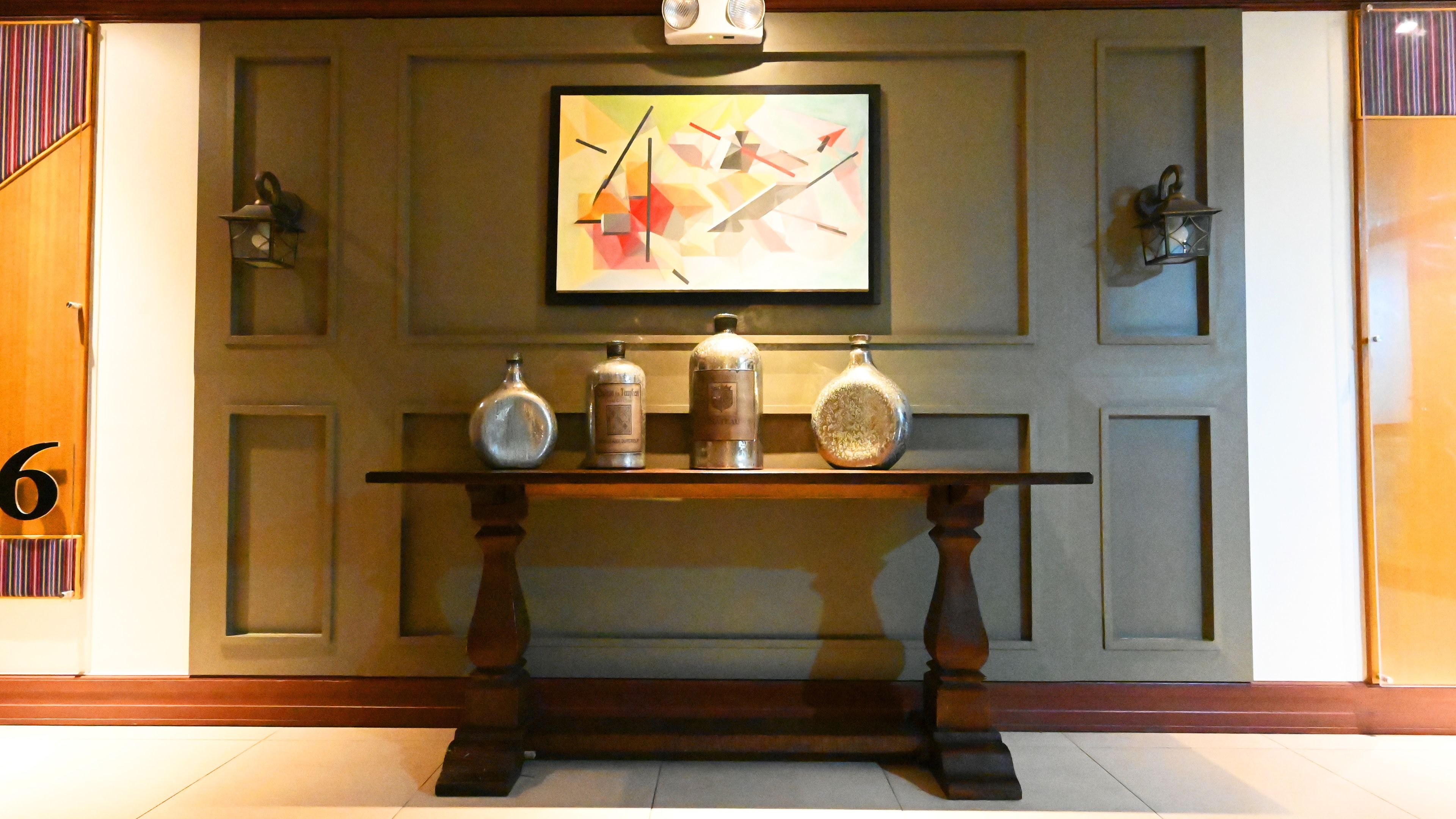 Een receptiegedeelte met een houten console tafel waarop drie flessen, een lijst met een abstract schilderij en twee wandlampen staan.