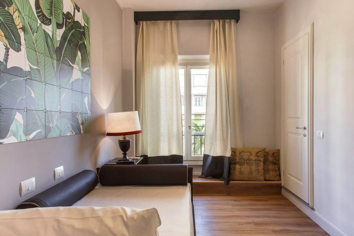 A hotel room with a bed, a nightstand, a lamp, a window with curtains, and a view of the outside.