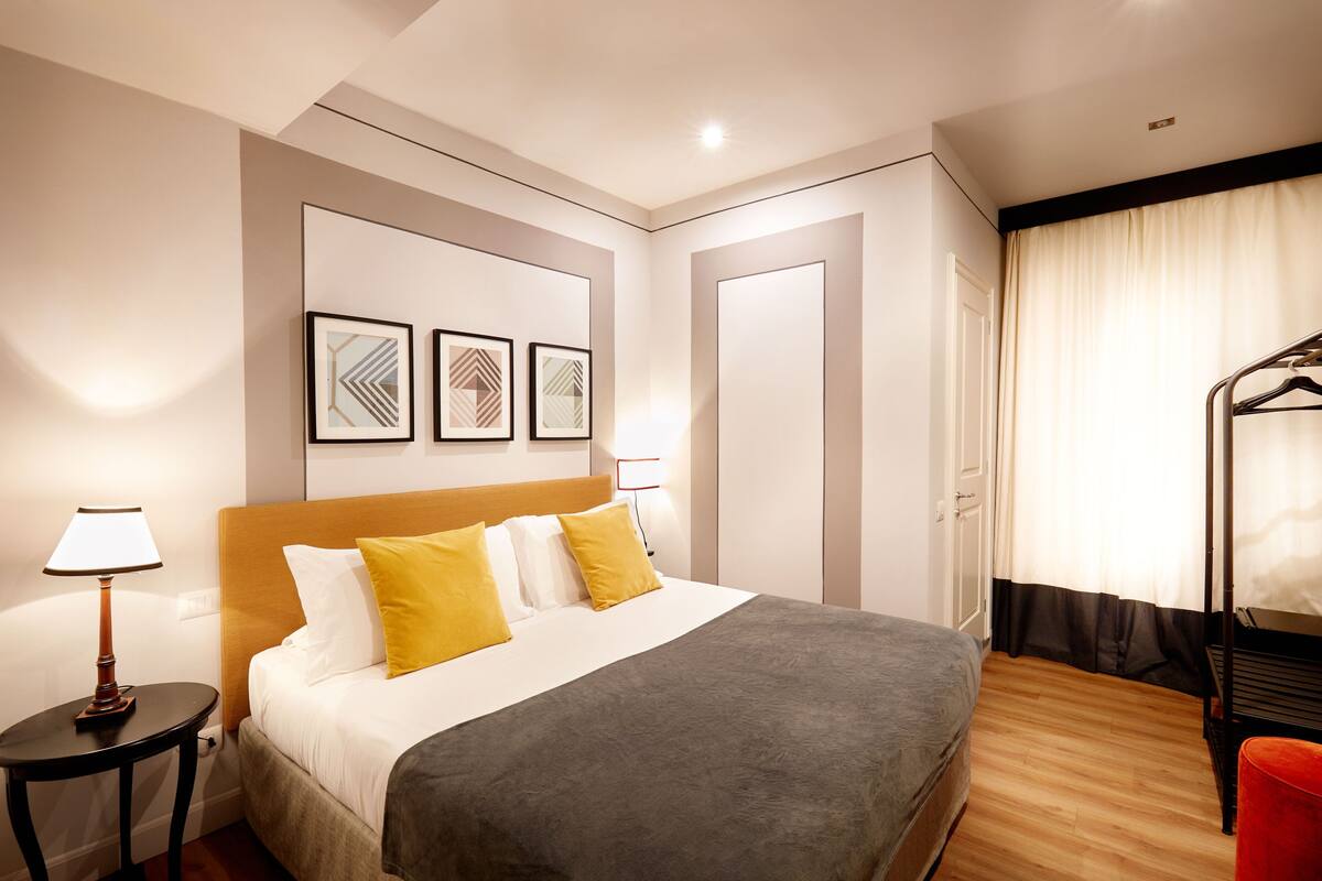 A hotel room with a bed, a side table, a lamp, a coat rack, and a door.