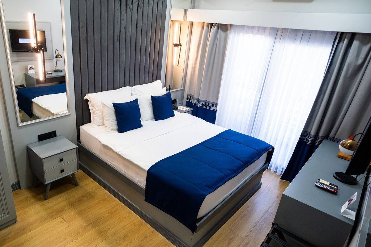 A modern hotel room with a large bed, a bedside table, a mirror, and a wardrobe.