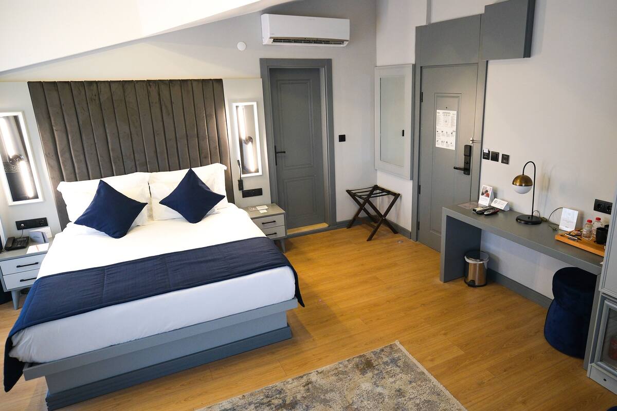 A modern hotel room with a large bed, bedside tables, a desk with a lamp, a wardrobe, and a trash bin.