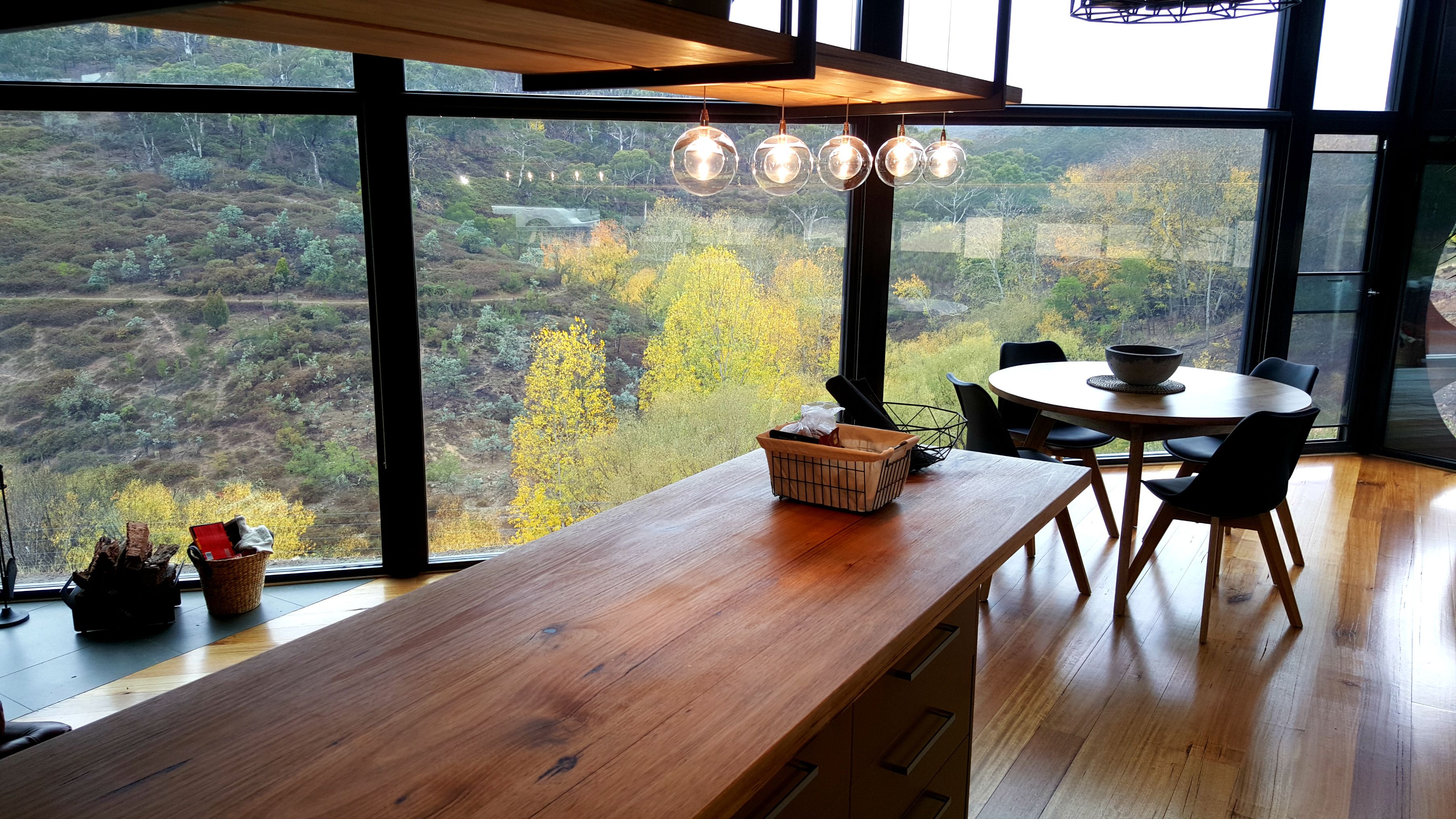 A modern kitchen with a wooden countertop, a dining area with a round table and chairs, and large windows offering a view of a forested landscape.