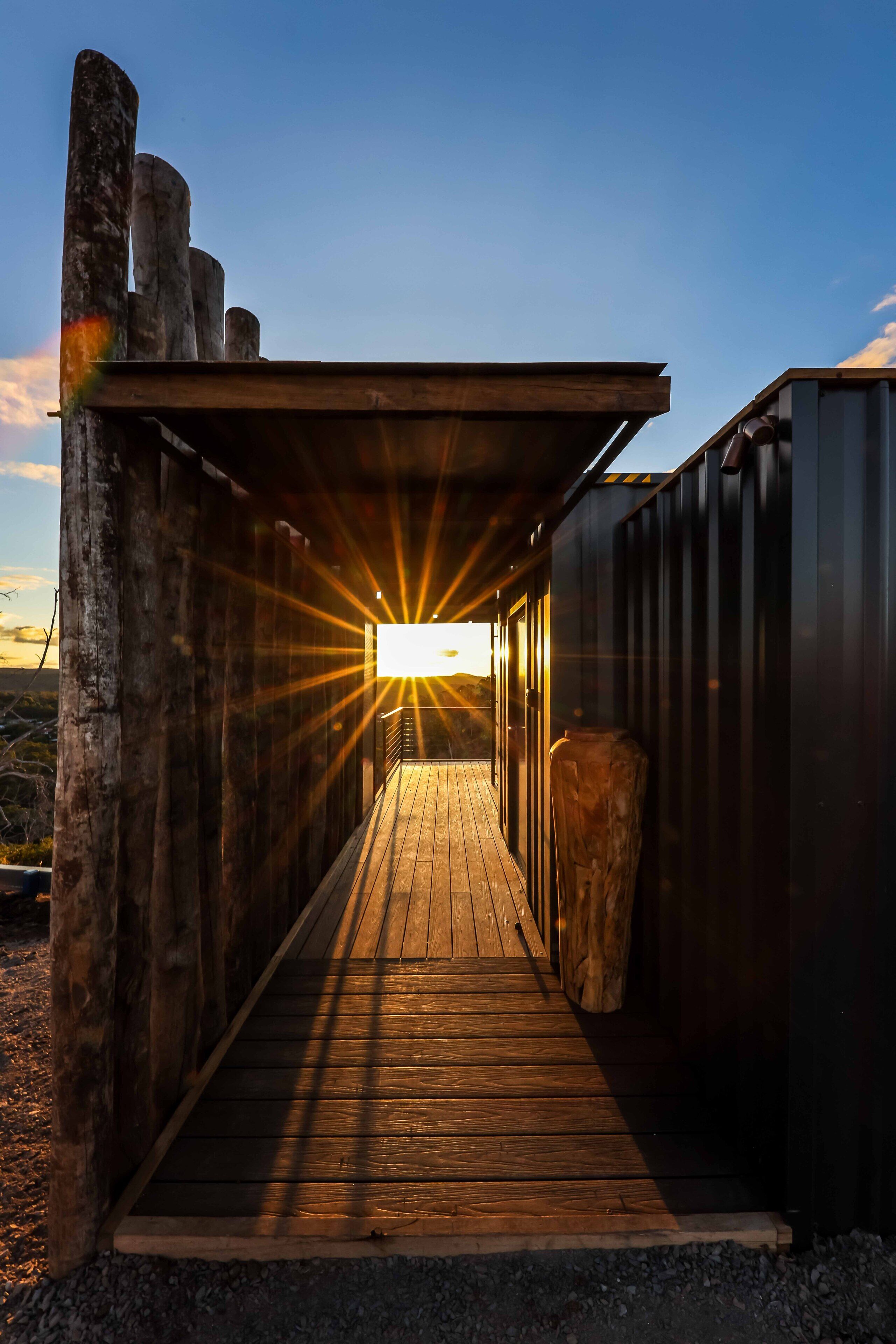 Un sendero de madera que lleva a un edificio, con el atardecer de fondo.
