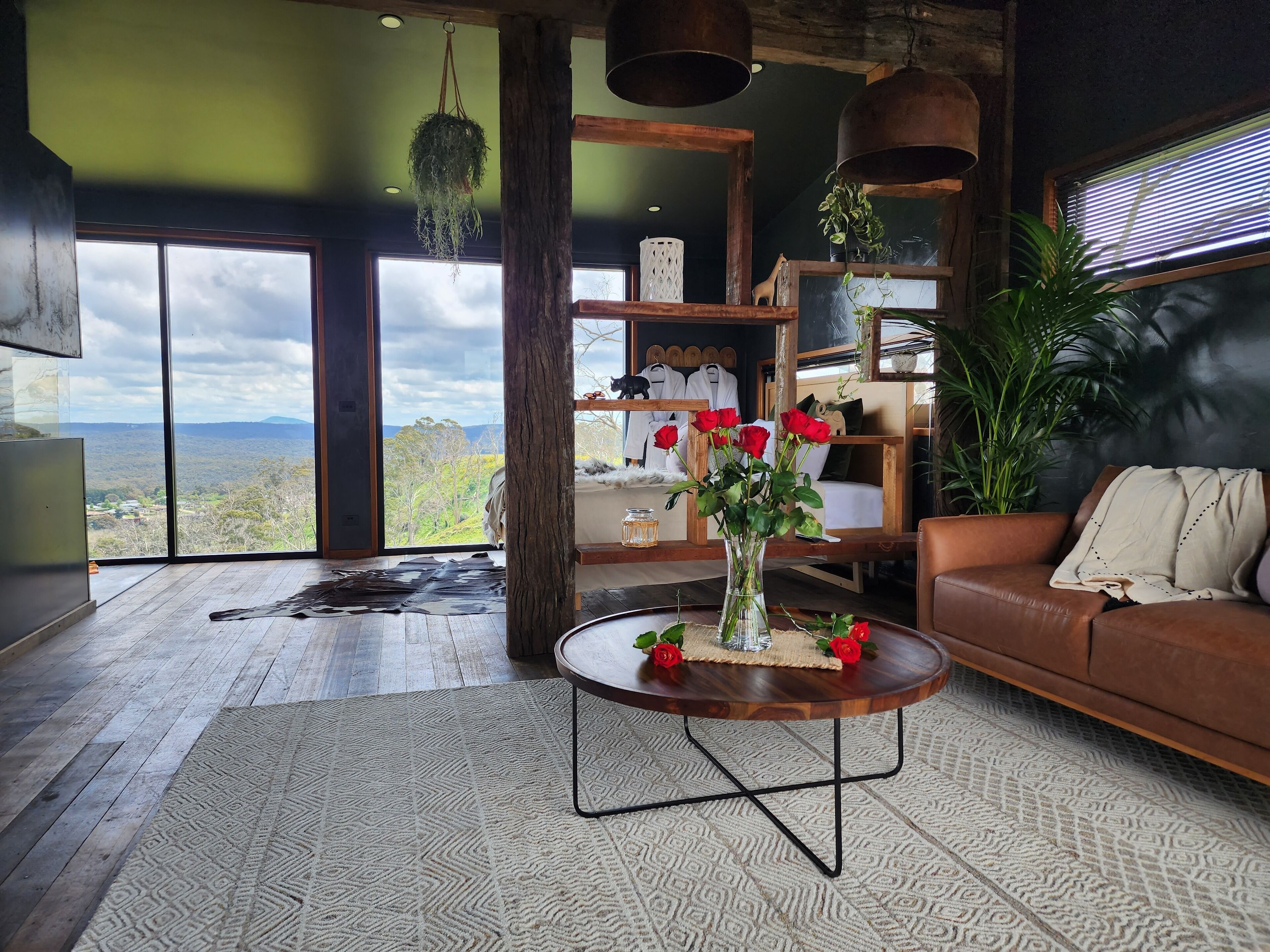 A spacious living area with a large window offering a view of the countryside, a round wooden coffee table with a vase of red roses, and a brown leather sofa.