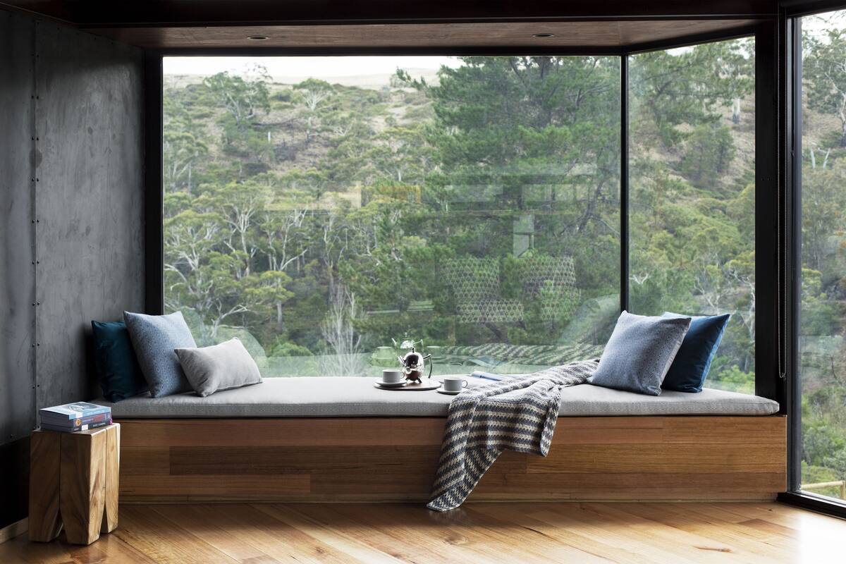 A modern room with a large window, a built-in bench with cushions, a small wooden stool, and a view of a forested area.