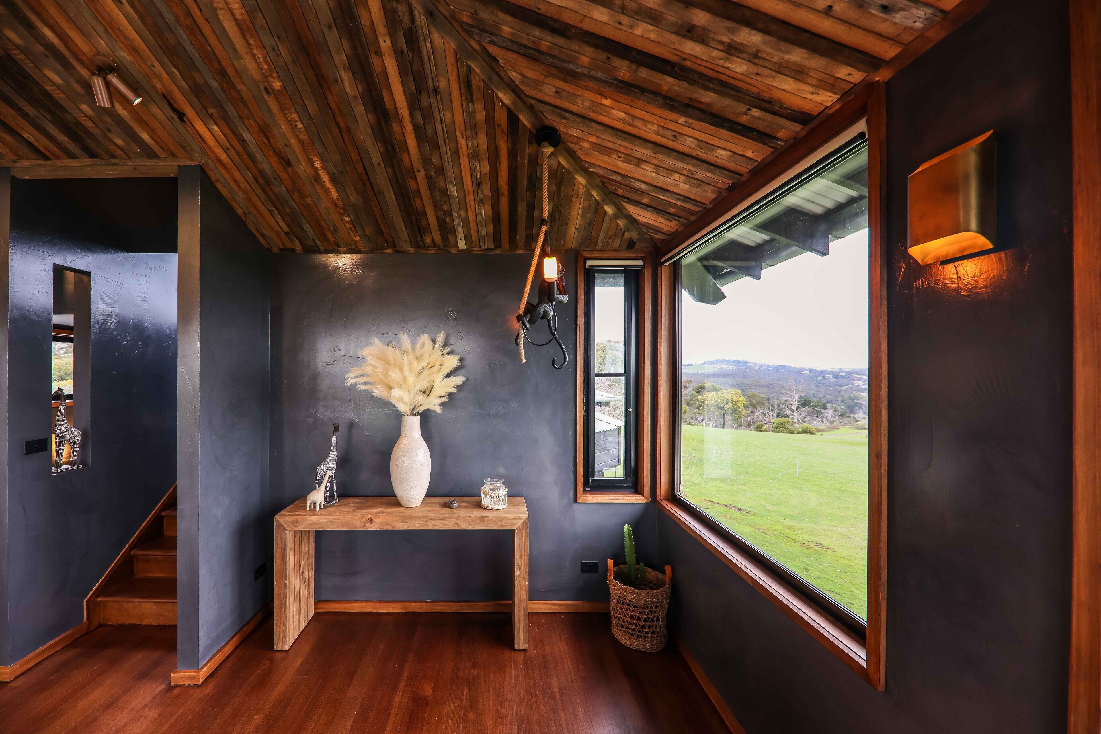 Een modern interieur met een houten plafond, een donkere wand, een houten console tafel met een vaas en beeldjes, een raam met uitzicht op de omgeving en een trap.