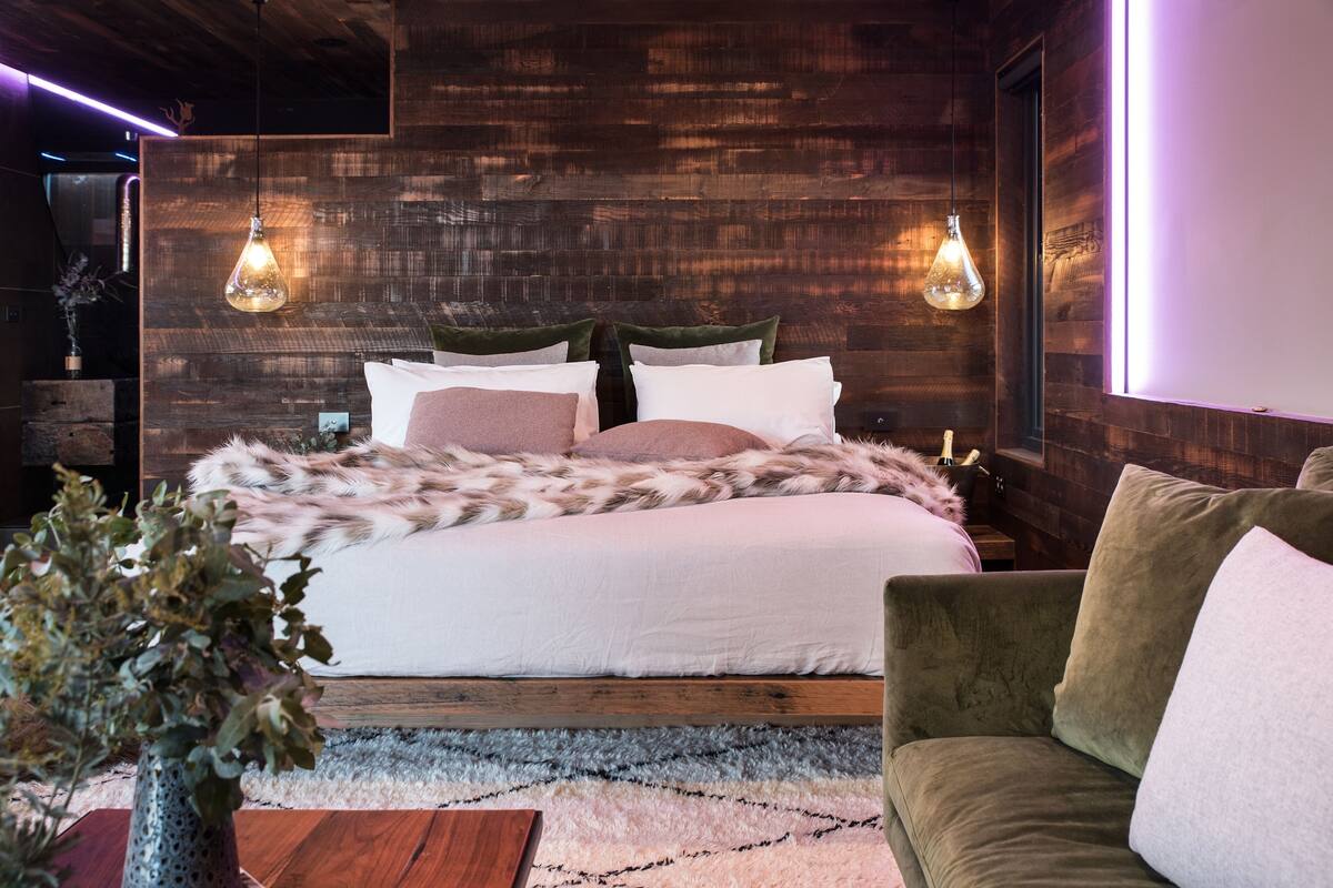 A modern bedroom with a wooden headboard, a large bed with multiple pillows, a green armchair, and two hanging light fixtures.