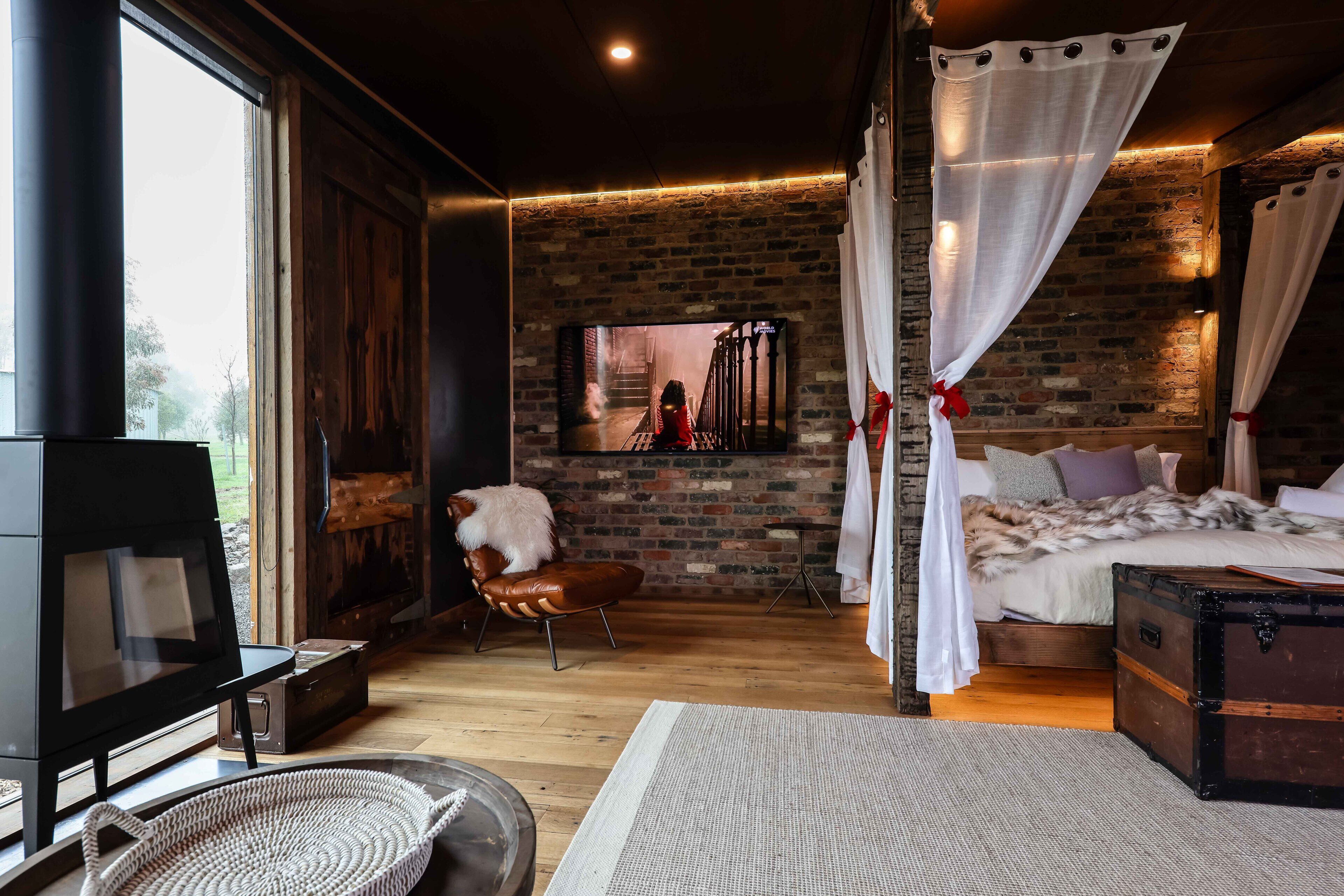 Una habitación acogedora con pared de ladrillo, un ventanal grande, una cama con ropa blanca, un cofre de madera y un televisor.