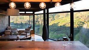 Un intérieur moderne avec un plafond en bois, de grandes fenêtres donnant sur une colline boisée, une table à manger avec deux verres à vin et un grand vase.