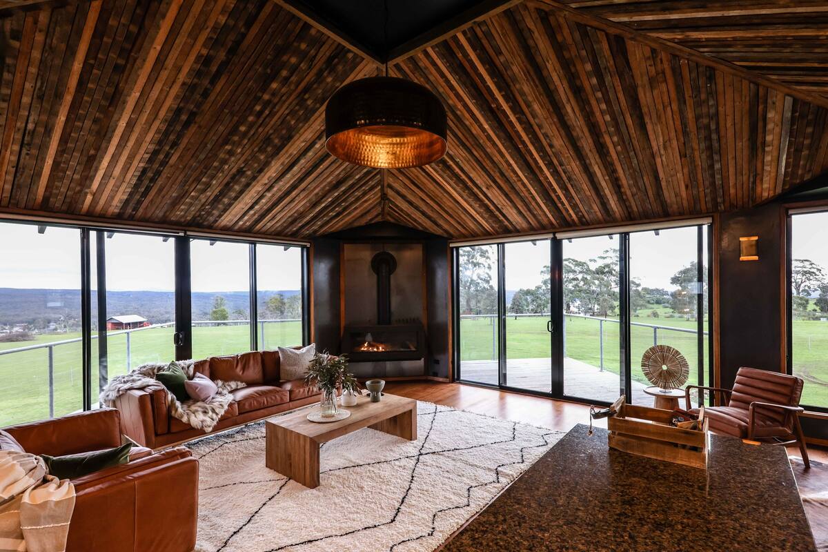 A spacious living room with a large fireplace, leather sofas, a coffee table, and a view of a green landscape through large windows.