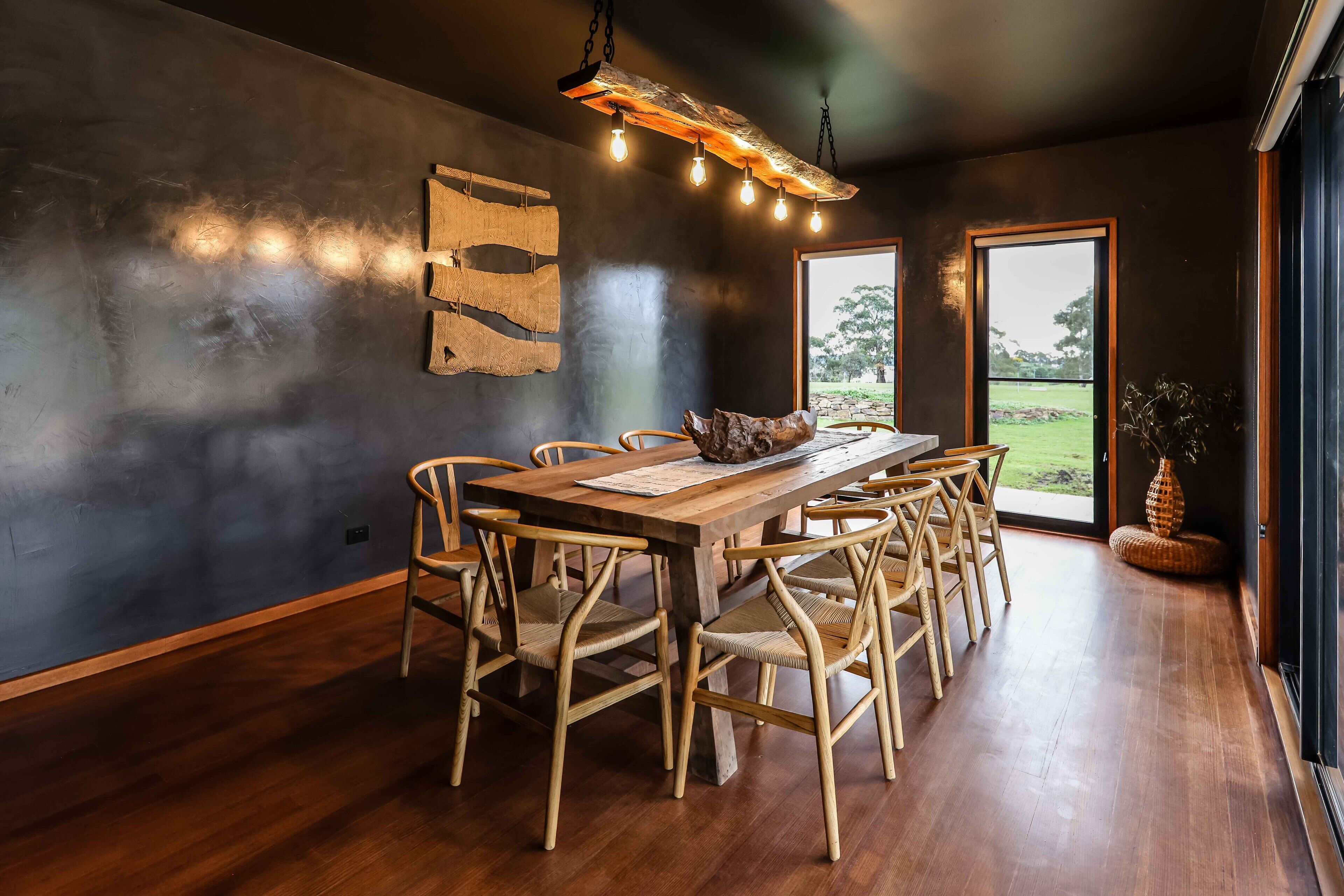 A dining area with a wooden table and chairs, a hanging light fixture, and a view of the outdoors through a large window.