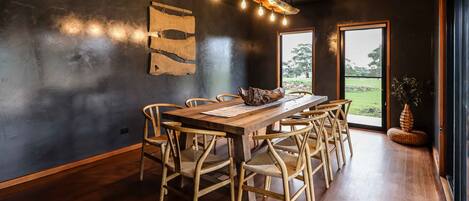 A dining area with a wooden table and chairs, a hanging light fixture, and a view of the outdoors through a large window.