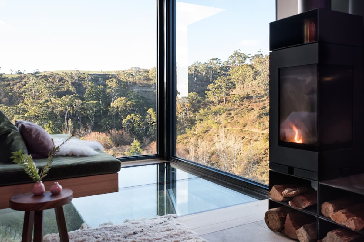 A modern living room with a glass floor, a sofa with cushions, a small table with a vase, and a wood-burning stove with logs.
