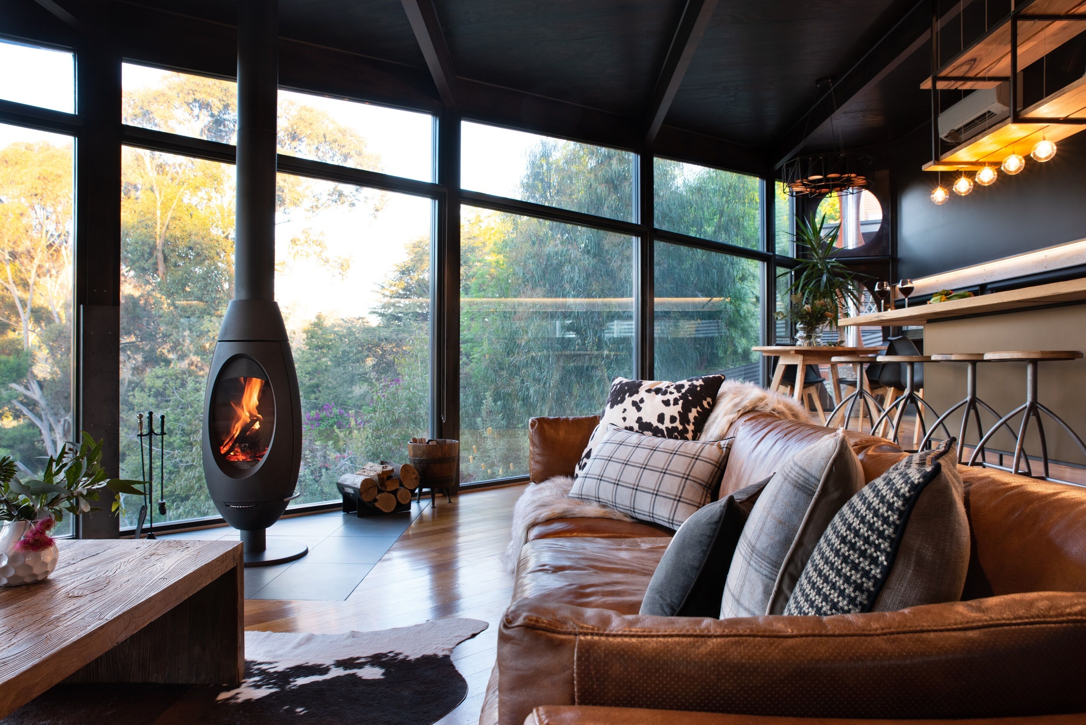Una sala de estar moderna con una chimenea grande, un sofá de cuero y vistas a árboles a través de amplios ventanales.