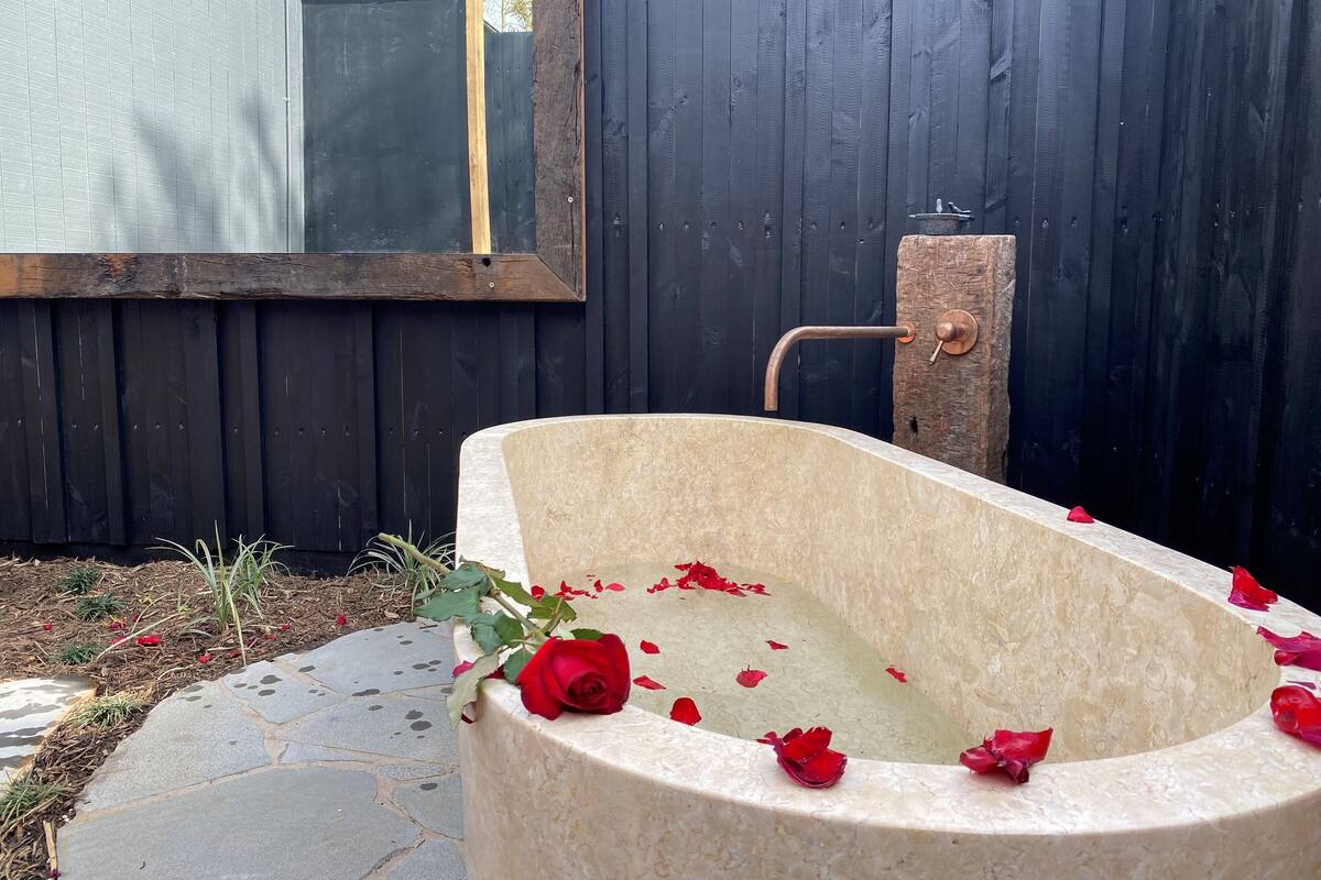 A stone bathtub with a rustic faucet, surrounded by rose petals, against a dark wooden fence.
