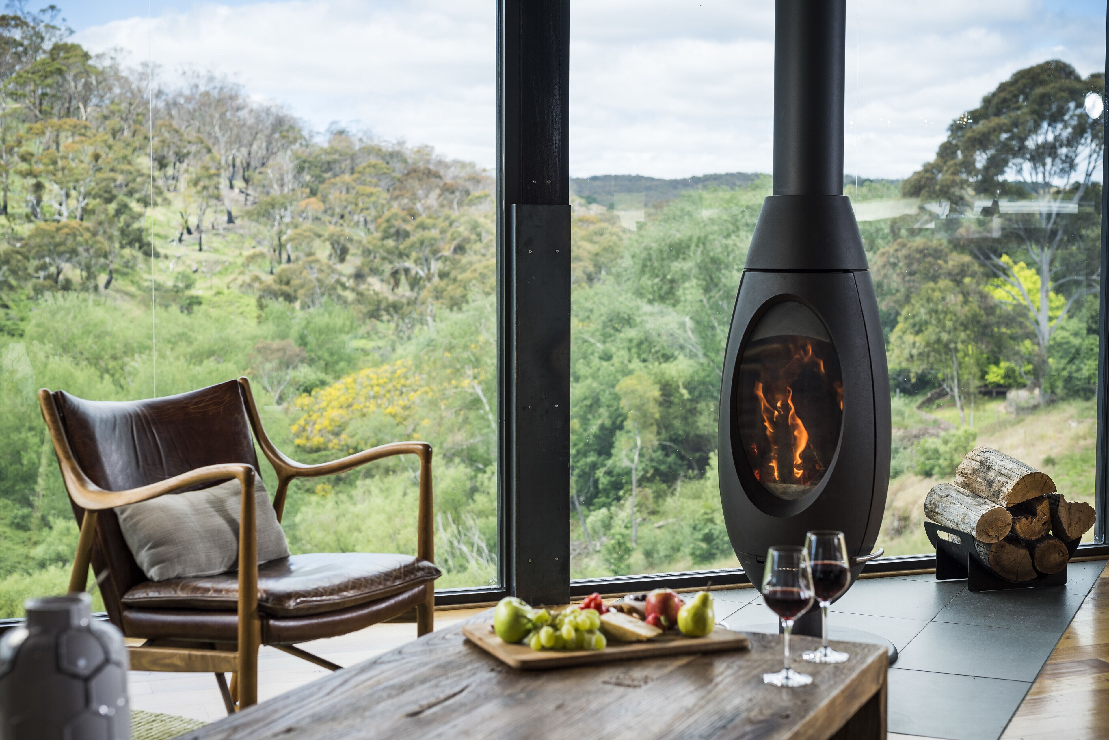 A cozy room with a large fireplace, a wooden table with fruit and wine glasses, and a comfortable chair.