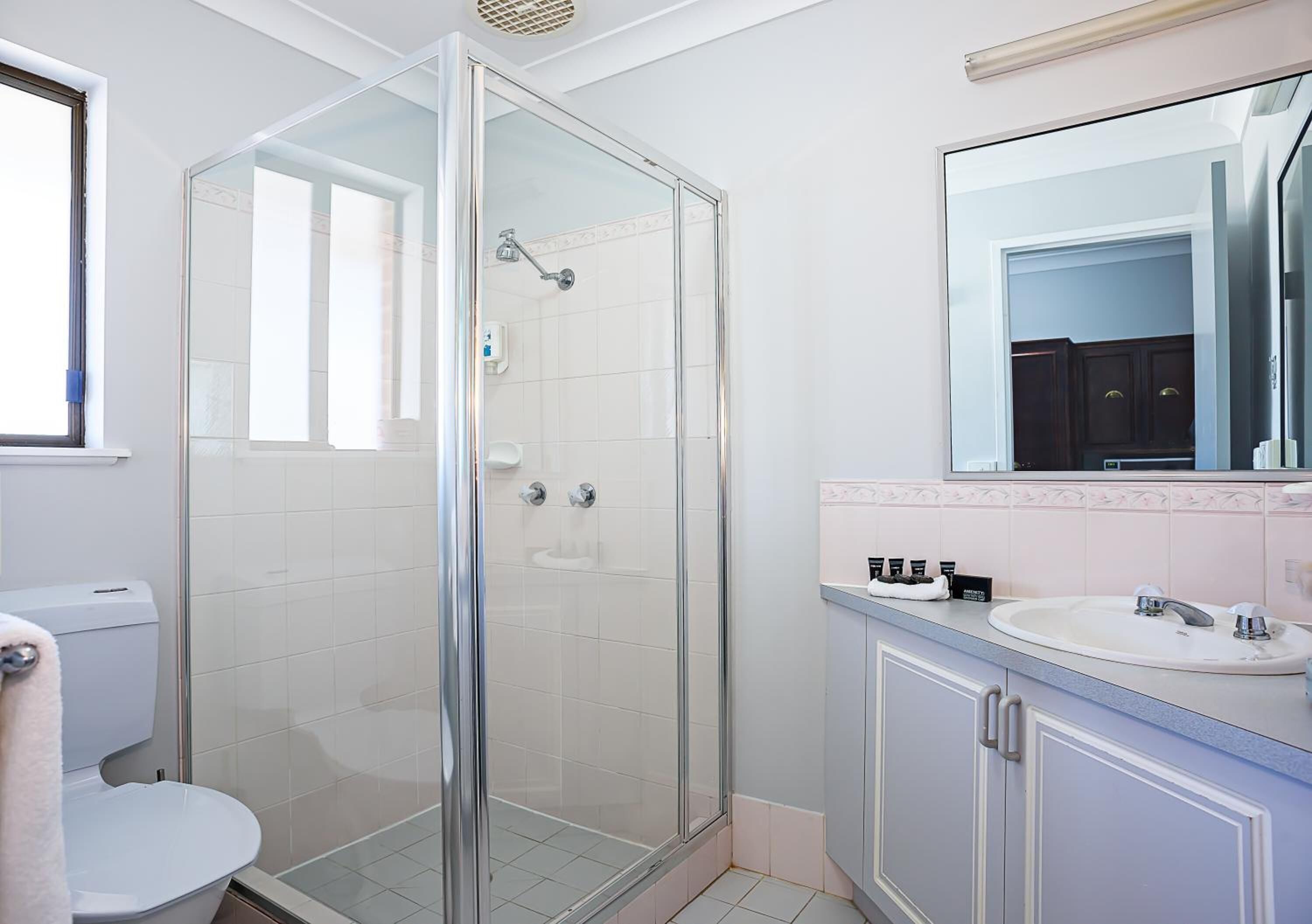 Un baño con ducha de vidrio, inodoro blanco, lavamanos con espejo y mueble bajo.