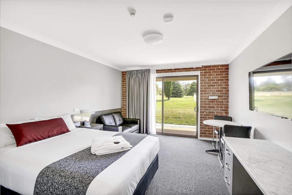 Habitación de hotel con cama, escritorio, silla y vista a un espacio verde a través de un ventanal.