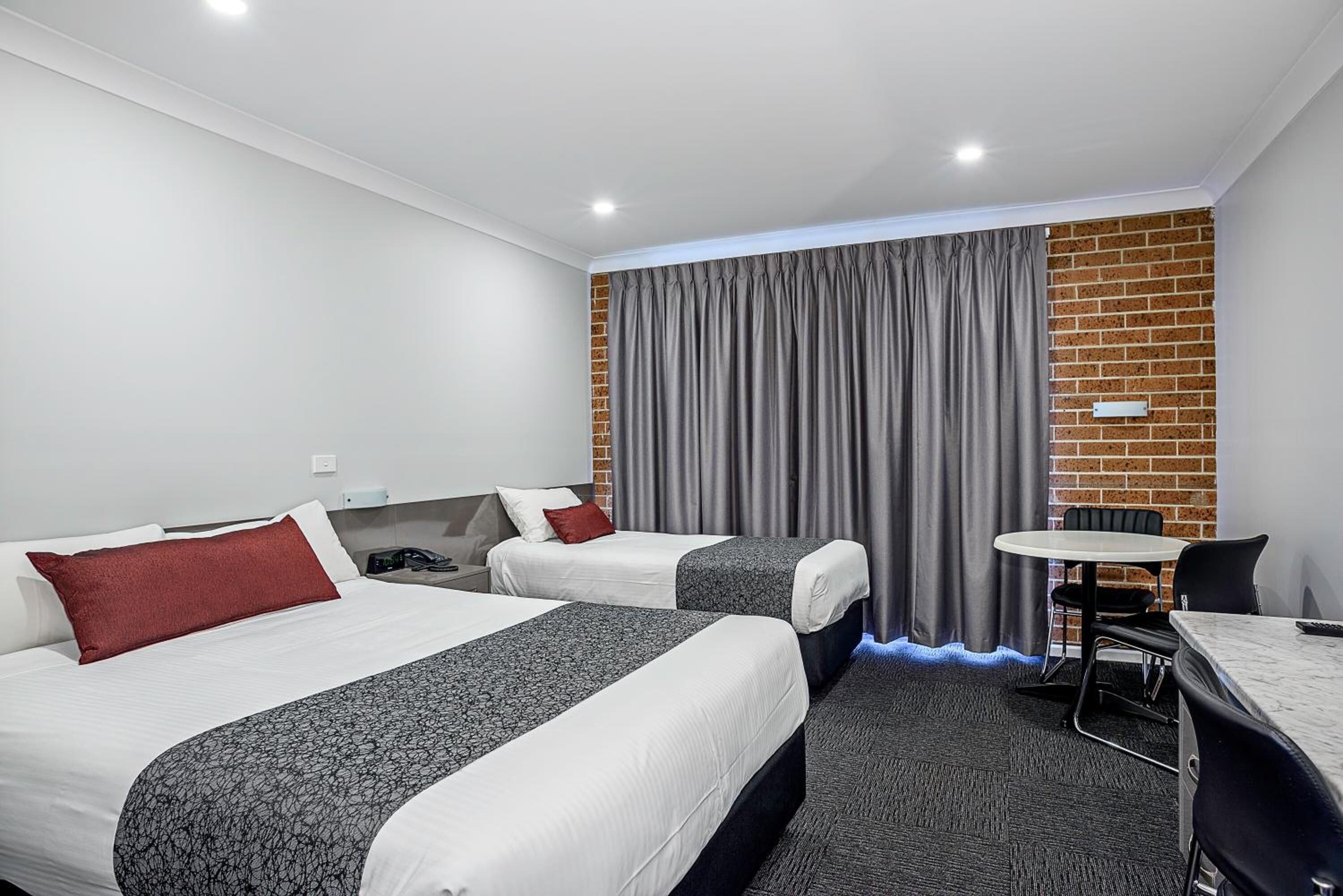 A hotel room with two beds, a small table, a chair, and a brick wall.
