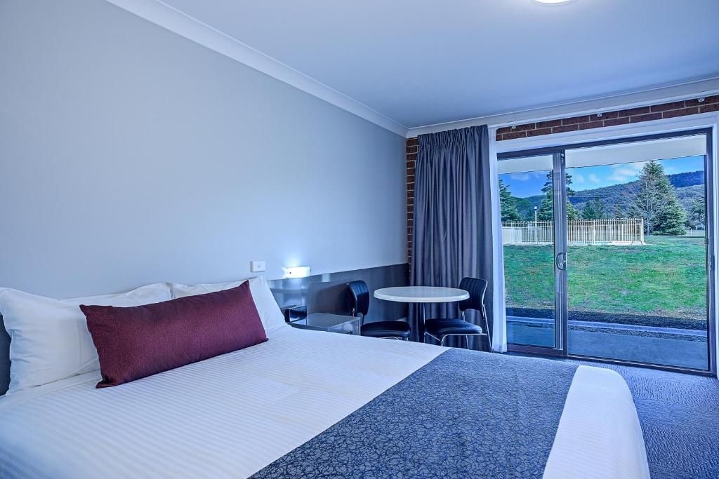 A hotel room with a bed, a small table with chairs, and a view of a grassy area through a glass door.