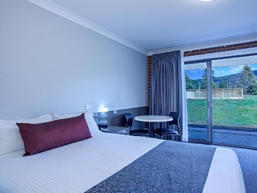 A hotel room with a bed, a small table with chairs, and a view of a grassy area through a glass door.