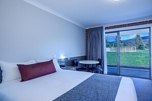 A hotel room with a bed, a small table with chairs, and a view of a grassy area through a glass door.