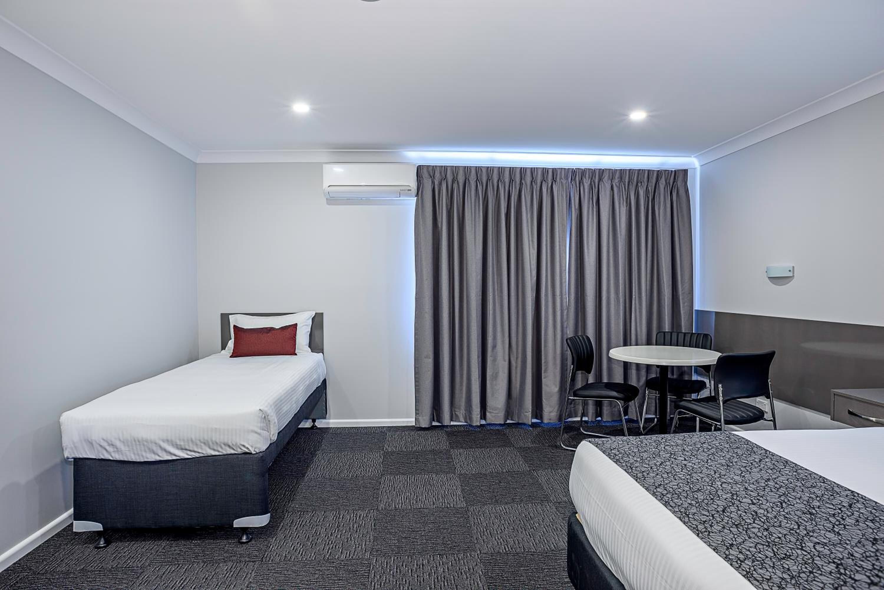 A hotel room with a single bed, a smaller bed, a desk with chairs, and a large window with curtains.