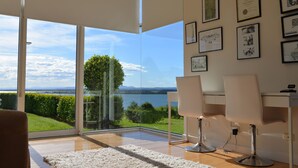 A modern room with a large glass door leading to a scenic view of a lake and mountains. There are two white chairs and a white desk with a computer. - Tintagel (Metung)