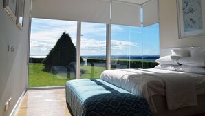 A bedroom with a large window, a bed with white bedding, and a view of a green landscape and water. - Tintagel (Metung)