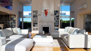 A spacious living room with two white sofas, a coffee table, and a fireplace. There are large windows providing natural light and a view of the outdoors. - Tintagel (Metung)