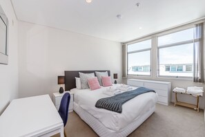 A modern hotel room with a large bed, a desk, and a view of the city through large windows.