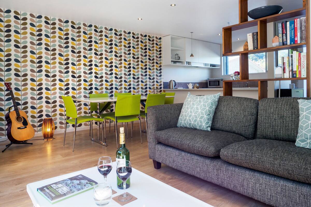 A modern living room with a grey sofa, green chairs, a dining table, and a bookshelf.