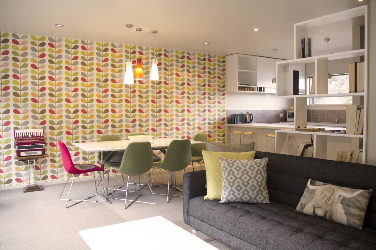 A modern living room with a grey sofa, colorful patterned wallpaper, and a dining area with green chairs.