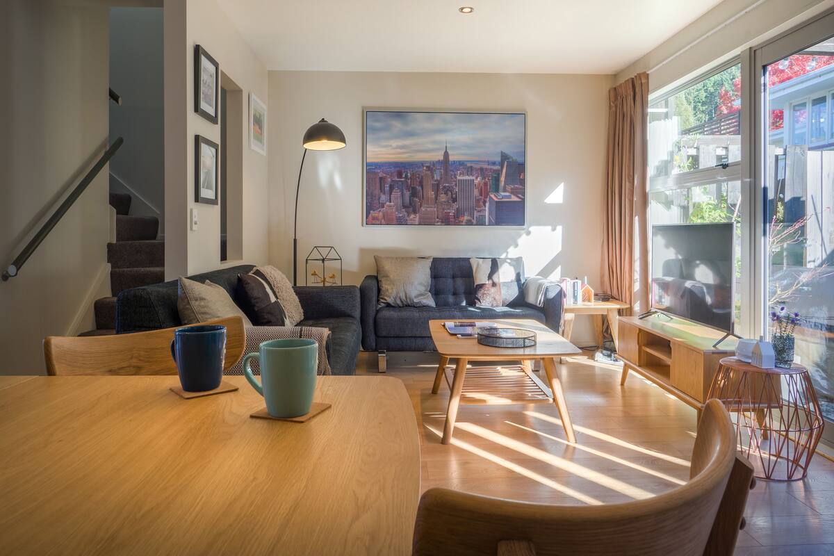 A modern living room with a sofa, coffee table, and a dining table. There is a staircase to the left and a large window with a cityscape view.