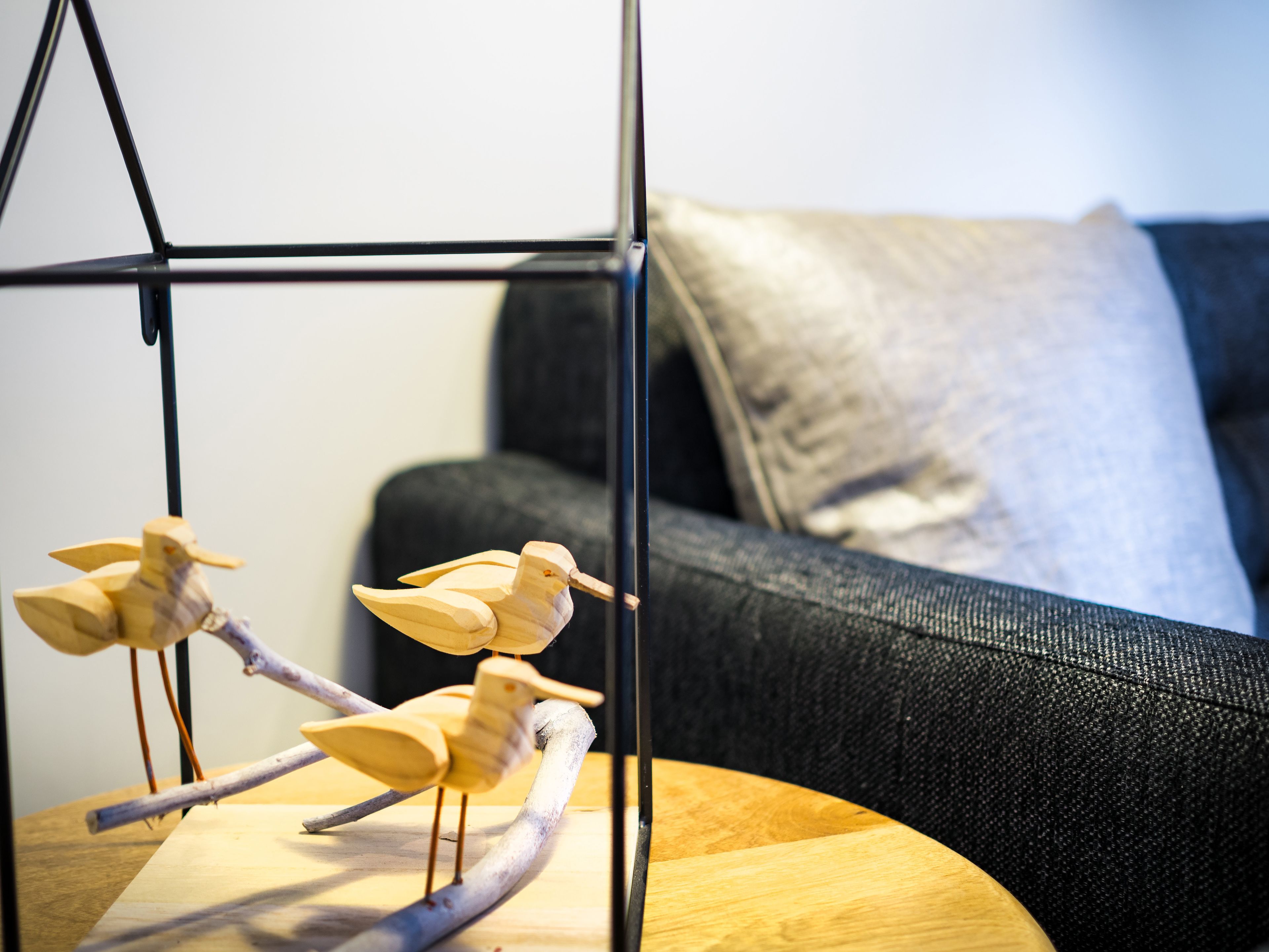 Una jaula de madera con ramas sobre una mesa de madera, un sofá negro con un cojín, y una pared de color claro al fondo.