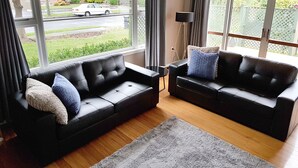 A modern living room with two black leather sofas, a grey rug, and large windows offering a view of a street and greenery. - Outdoor Living in Christchurch (Christchurch)