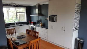 A modern kitchen with white cabinets, a dining table with chairs, a stove, a microwave, and a window with a view of greenery. - Outdoor Living in Christchurch (Christchurch)