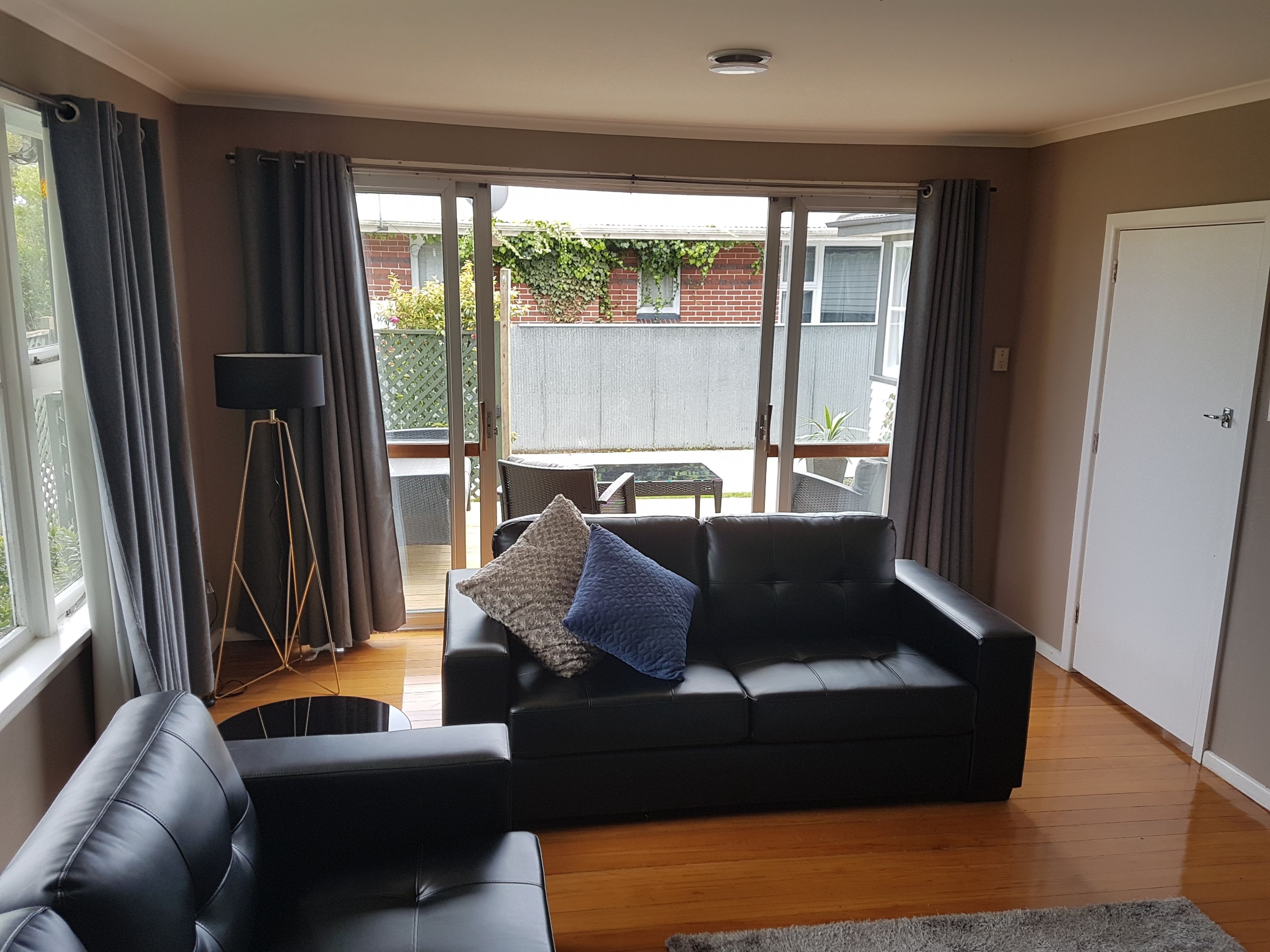 A living room with a black leather sofa, a floor lamp, and a view of the backyard through glass doors.