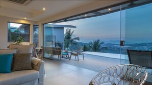 A modern living room with a sofa, a dining table, and chairs. There is a large glass window offering a view of the city and mountains. - Villa Lanta at Comoon Boutique Villas (Koh Samui)