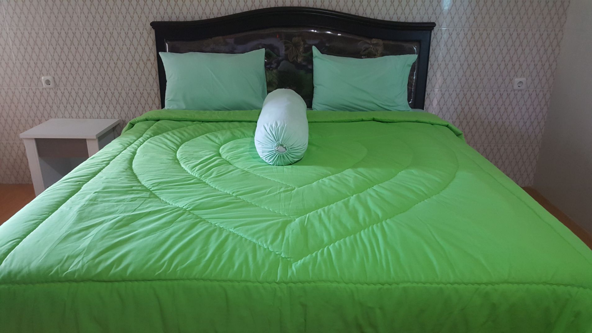 A bed with a green quilt and a white pillow, a wooden headboard, and a small bedside table.