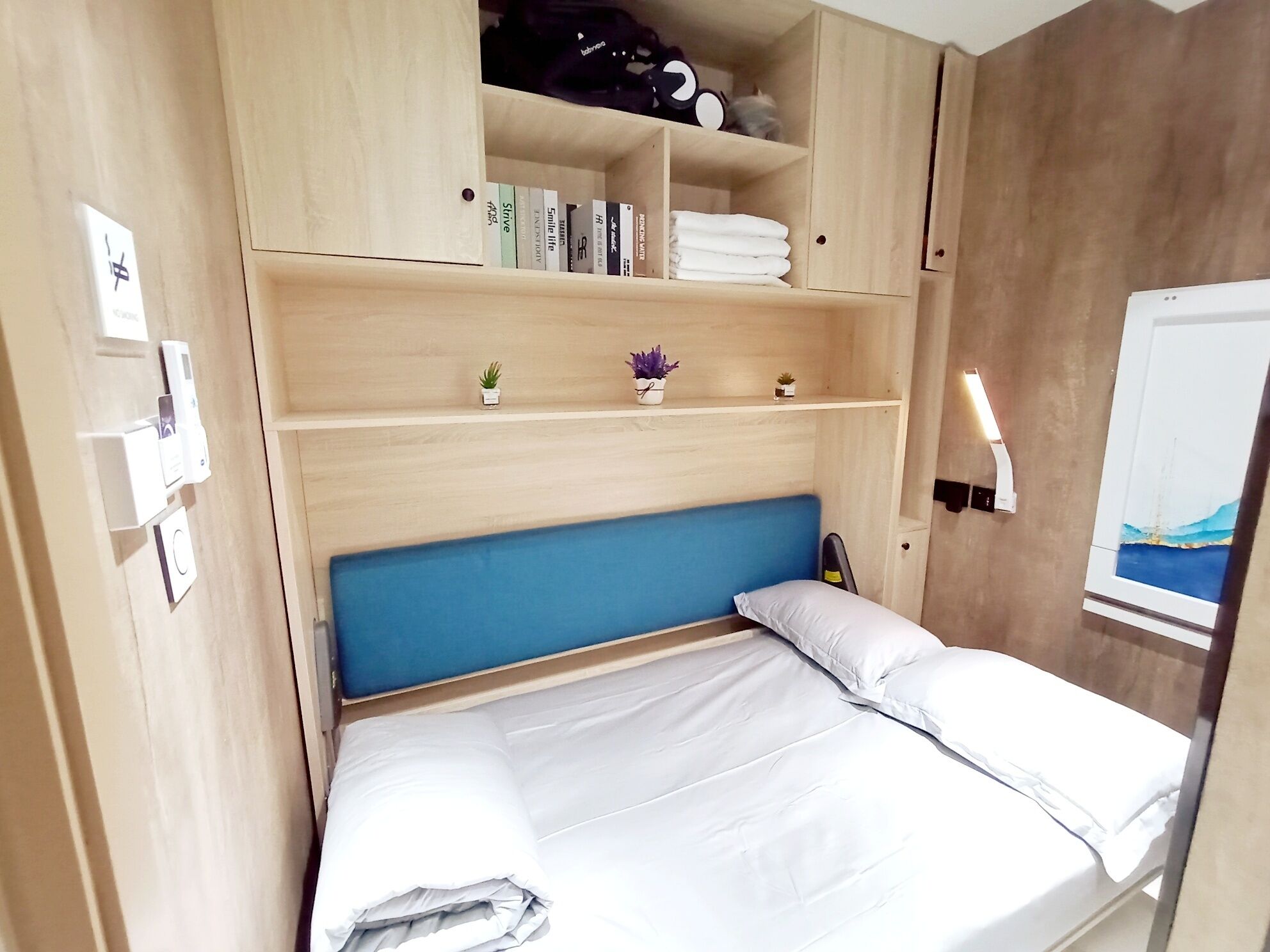 A compact room with a bed, a shelf with books, a small plant, and a whiteboard.